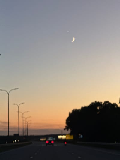 The image depicts a highway scene at dusk in Piņķi, Latvia. The sky transitions from a pale blue at the top to a warm orange and yellow hue near the horizon. A crescent moon and a small, bright celestial body are visible in the upper portion of the sky. Along the left side of the highway, a series of streetlights are present, with one prominently in focus and others receding into the distance. A dark silhouette of trees forms the right boundary of the scene. On the highway, there are several vehicles. One car with illuminated red taillights is visible in the foreground, traveling away from the viewer. Other vehicles, with their headlights and taillights illuminated, are further down the road. The road surface is dark asphalt with white lane markings. The overall impression is that of a twilight drive on a major road.
