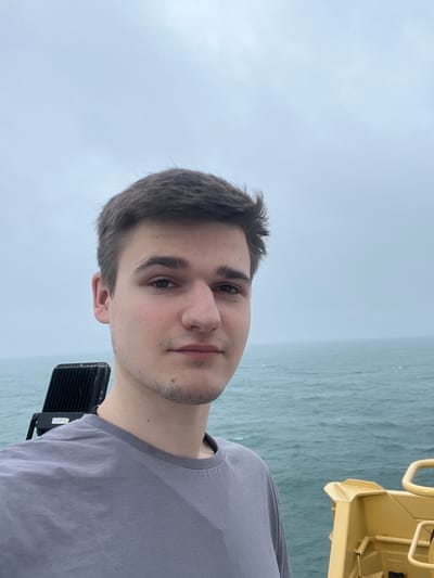 A young adult male, approximately 18-25 years old, with short dark brown hair, fair skin, a prominent nose, and light facial stubble on his chin and upper lip, is depicted facing the viewer with a neutral expression. He wears a plain grey crew-neck t-shirt. The scene is outdoors on what appears to be a vessel or platform at sea, consistent with a coastal location such as Bangor, United Kingdom, though specific identifying landmarks are not visible. Behind the male's left shoulder, a black, rectangular industrial floodlight with a finned heat sink is mounted on a structure. To the male's right and extending into the background, a choppy, greenish-blue sea with visible waves dominates the mid-ground. The sky above is entirely overcast, a uniform light grey that blends into a hazy horizon with the sea. In the lower right corner, a section of a yellow metallic structure, possibly a railing or equipment component, is visible.