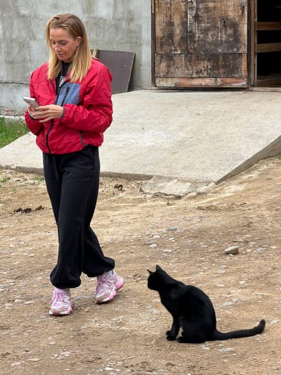 A woman stands in a dirt area next to a concrete path. She is wearing a red jacket, black pants, and pink and white sneakers. She is looking down at her phone, which she is holding in her left hand. A black cat is walking on the dirt to her right. In the background, there is an old wooden door and a concrete wall. The scene appears to be outdoors, possibly in a rural or industrial area. The lighting suggests it is daytime. The woman's expression is focused on her phone, and the cat seems indifferent to her presence. There is no visible text in the image.