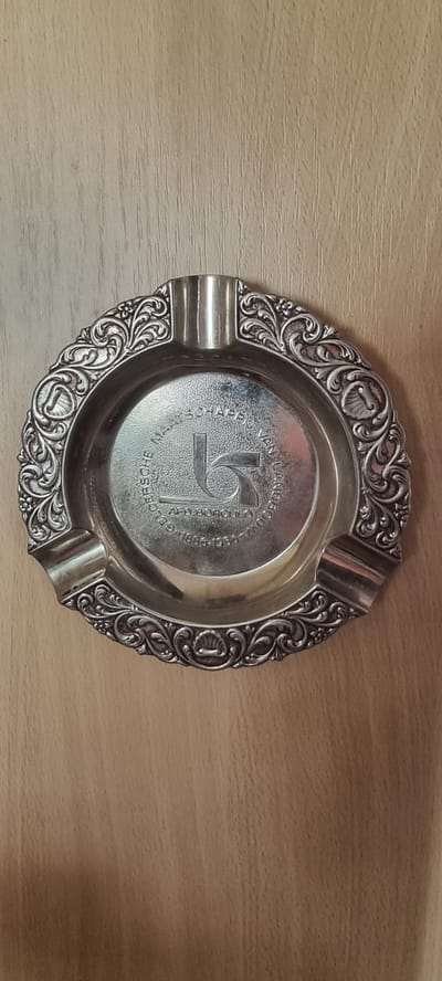 An ornate, silver-plated ashtray with decorative scrollwork and shell motifs rests on a light brown, wood-grained surface. The ashtray features four indentations for cigarettes and a circular depression in the center. Engraved within the center depression is a stylized "LS" logo, and around the outer edge of the depression are the words "GEVERSCHE MAATSCHAPPIJ VAN .." followed by "AFD. BORCULO" and the dates "1886-1986". The ashtray is centrally placed within the frame, casting a subtle shadow. The background is a plain wooden surface, with visible vertical grain. There are no people or active events depicted in the image.