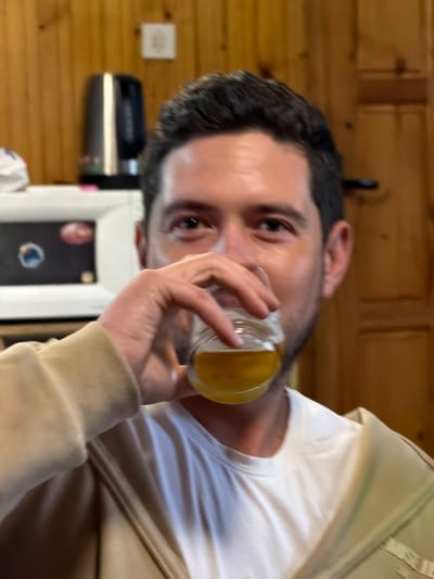 A man is drinking from a glass of yellow liquid. He has short dark hair, fair skin, and is wearing a white t-shirt and beige jacket. The background is a wood-paneled wall and a white object with a dark screen. The location is Lilkovo, Bulgaria.