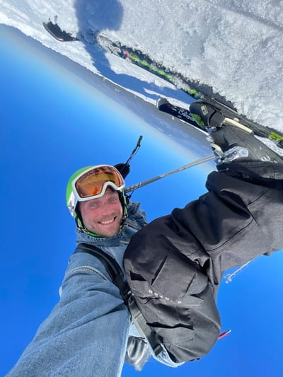 An upward-angled image captures a smiling male individual on skis in a snow-covered landscape under a clear blue sky, identified as Jahorina, Bosnia and Herzegovina. The male, with light skin and a short beard, is wearing a bright green ski helmet with orange-tinted goggles, a light blue denim jacket, and black ski pants with "BURTON" visible on the left thigh. Black backpack straps are visible over his shoulders. He holds a silver ski pole, marked "LEKI FREEZE," in his gloved right hand. In the upper right quadrant, two skis are partially embedded in the textured white snow; one ski exhibits "Elan" branding and a black binding attached to a black ski boot. The dominant background is a clear, vibrant blue sky. Shadows suggest bright sunlight.