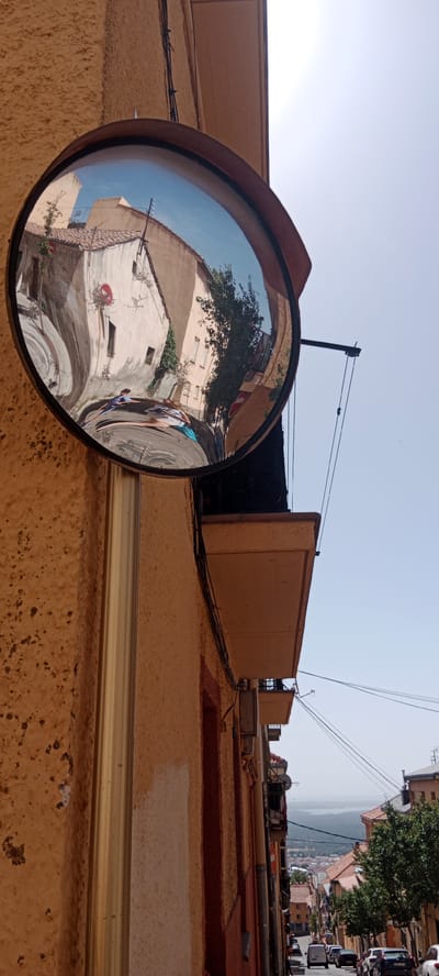 This media file is a photograph taken outdoors in San Lorenzo de El Escorial, Spain. The image features a convex mirror mounted on a textured, ochre-colored building. The mirror reflects a scene of a narrow street lined with buildings, with two people walking. The buildings appear to be old, with tiled roofs and light-colored facades. One building in the reflection has a tree growing on its side, and a red circular sign with a white line through it is visible. The sky in the reflection is clear and blue, suggesting a sunny day.

Beyond the immediate reflection, the photograph shows a balcony extending from the building where the mirror is mounted. The balcony has a light-colored surface and is supported by metal brackets. Electrical wires are visible running along the side of the building and across the sky. Further in the distance, the view opens up to a vista of a town or village nestled in a valley, with more buildings and trees. A body of water and hills can be seen on the horizon. Several cars are parked or driving down the sloping street in the background.

The time of day appears to be daytime, given the bright sunlight. The weather seems to be clear and fair. The overall impression is one of a quiet, sun-drenched European street scene. No text is clearly legible in the image.