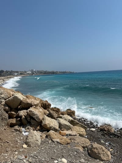 The image shows a rocky coastline with the turquoise sea in Kissonerga Community, Cyprus. In the foreground, there are large, weathered rocks and pebbles, with some scattered debris. The sea is visible in the midground, with waves crashing against the shore, creating white foam. In the background, a town with white buildings lines the coast, and the sky is clear and blue. The scene depicts a natural coastal landscape with signs of human presence and environmental impact from debris.