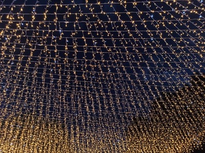 The image shows a low-angle view of a large canopy of string lights at night. The lights are a warm yellow color, with some interspersed blue lights. They are arranged in a grid-like pattern, with strings of lights hanging down from horizontal wires. The background is a dark night sky, suggesting the time is after sunset. The lighting creates a festive and magical atmosphere. There are no people or specific objects visible beyond the lights and their supporting wires. The scene appears to be an outdoor installation, possibly for a holiday celebration or a decorative display in a public space. The overall impression is one of wonder and enchantment, illuminated by the multitude of tiny lights.