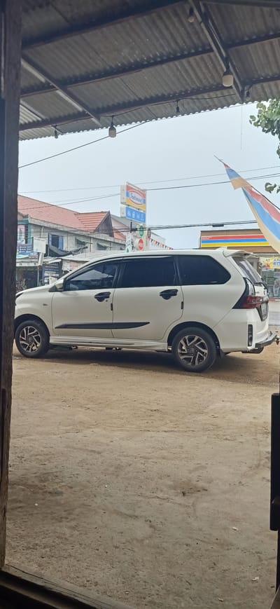 A white minivan is parked on a dirt lot in front of buildings in Uteungeulinggang, Indonesia. The minivan is facing to the right, with its doors closed and windows tinted. The lot is dusty and uneven, with some scattered debris. In the background, there are several buildings, including a store with an "Indomaret" sign. A corrugated metal roof extends from a structure in the foreground, supported by wooden posts. The sky is overcast.