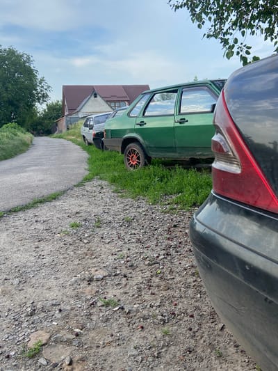 The image depicts a quiet street scene in Mala Vilshanka, Ukraine, with a focus on parked cars and the surrounding environment. In the foreground, the rear quarter panel of a dark-colored car is visible, hinting at its presence. To the left of this car, a row of other vehicles is parked along a grassy verge bordering a paved road. Among these, a distinct green car with orange-rimmed wheels stands out. Further down the road, a light-colored car and another vehicle are partially visible. The road itself is a paved surface that curves gently to the left, leading towards a residential area with a prominent house featuring a dark-tiled roof. Trees and foliage flank the road and residential property, suggesting a rural or semi-rural setting. The sky above is partly cloudy, with patches of blue sky visible through the clouds. The ground is a mixture of gravel and dirt, with scattered small red berries or fruits on the surface. In the distance, on the left side of the road, a small group of people can be seen on bicycles, adding a touch of activity to the otherwise still scene.