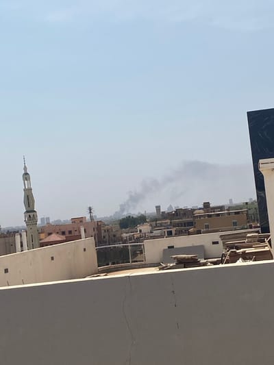 The image shows a view of a cityscape in Omdurman, Sudan, taken from a rooftop. The scene is dominated by a large, gray building with a flat roof. The building appears to be constructed from concrete and has a crack running down the right side. In the background, a cityscape of brown buildings is visible, with some buildings having multiple stories. A large plume of smoke is rising above the city, suggesting a possible fire or industrial activity. A single palm tree is visible in the middle distance. A tall, slender structure with a pointed top, possibly a minaret, is located in the foreground. There are no people visible in the image.