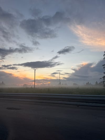The image depicts a serene, foggy landscape at dusk or dawn, viewed from the side of a road. The foreground is dominated by the dark asphalt of the road, with a gray guardrail separating it from a field. The field is shrouded in a soft mist, with silhouetted trees and vegetation faintly visible in the distance. Two utility poles with overhead wires are prominent features in the middle ground. The sky above is a canvas of soft hues, with a mix of gray, blue, and peach-colored clouds, suggesting the last light of day or the first light of dawn. The overall impression is one of tranquility and natural beauty, with a subtle hint of human infrastructure. The location in the city of Kapan', Russia, is not discernible from the visual elements present in the image.