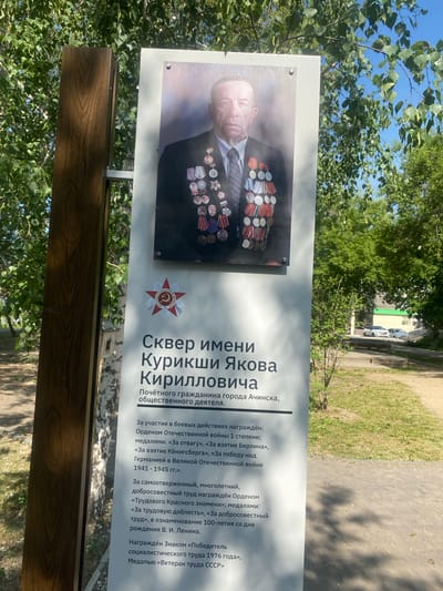 The image shows a memorial plaque with a portrait of an elderly man adorned with numerous medals. The plaque is situated outdoors, with trees and a blue sky visible in the background. The text on the plaque indicates that it is dedicated to Skver imeni Kurikshi Yakov Kirillovich and mentions his status as an "Honorary Citizen of the city of Achinsk, public figure." The text also lists his military decorations and awards for labor. The plaque is mounted on a white board, attached to a wooden post, and positioned near a paved walkway in what appears to be a park or public square. The overall scene suggests a commemorative display honoring a local hero in Achinsk, Russia.