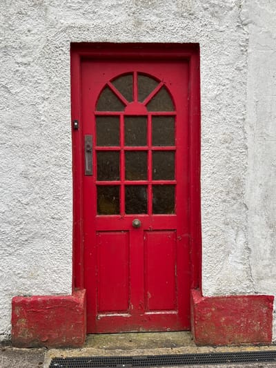 The image shows a red wooden door set into a white stucco wall. The door has a decorative window with a fan-shaped leading above a grid of nine square panes. The glass in the panes is textured and dark. There is a brass letterbox and a small doorbell button below the window. To the right of the door, there is a section of the door with two raised panels. The door appears to be old, with some peeling paint. The surrounding wall is rough and textured. Red painted stone blocks are visible at the top right and bottom right corners of the door frame, suggesting it is recessed into the wall. To the far right, a black metal grate is visible. The scene suggests an exterior view of a building in Mountcharles, Ireland. There are no people visible, and no specific activity is taking place. The lighting appears to be natural daylight.