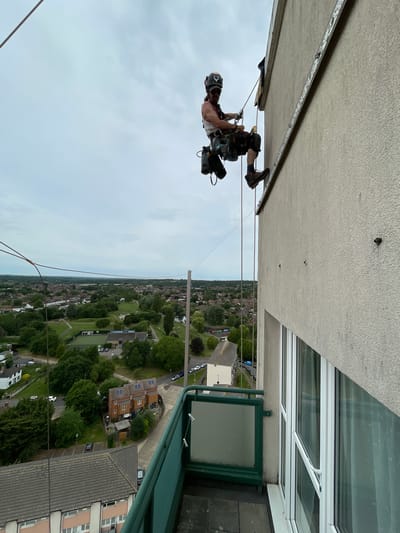 A worker rappels down the side of a building in Hertsmere, United Kingdom. The worker is suspended by ropes attached to his harness and wears a helmet. He is equipped with tools and gear attached to his harness. The building is multi-story, with windows and a balcony. The surrounding area is residential, with houses, trees, and green spaces. The sky is overcast.