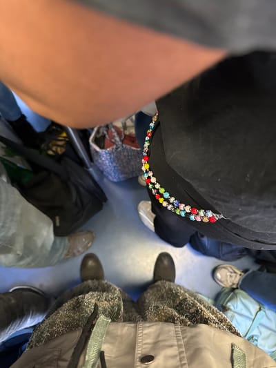 This high-angle image captures the lower bodies, feet, and belongings of several individuals in a crowded indoor setting, possibly public transport in Bourg-la-Reine, France. The floor is a light blue-grey color.
In the center, a person wearing black clothing has a decorative, colorful beaded accessory, featuring red, blue, green, yellow, pink, and clear beads, draped from their waist or hip. This individual wears light-colored shoes. To their right, another person is seen with dark shoes or boots and a grey puffy jacket over a textured or furry garment.
Further into the background on the right, a person in light blue jeans wears light brown or beige shoes. Another individual with dark shoes and grey or blue jeans is partially visible. On the floor between the people are several bags: a light blue or mint green duffel bag or backpack, a black backpack, and a smaller patterned fabric bag. The close proximity of the subjects and their belongings suggests a confined space, like a bus or train carriage. No specific activity beyond gathering or traveling is depicted, and no text is visible.