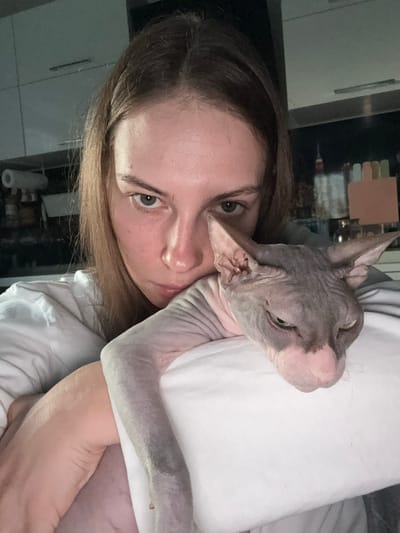 A close-up, selfie-style photograph shows a young woman with light brown hair and light-colored eyes, wearing a white t-shirt, holding a Sphynx cat. The woman's head is angled towards the camera, and she is looking directly at the viewer with a neutral expression. The cat, which is grey and pink with prominent wrinkles, is resting on the woman's chest and shoulder. Its head is turned away from the camera, with its eyes partially closed. The background appears to be an indoor kitchen environment, with cabinets and shelving visible, suggesting the location is a residence in the city of Novaya Ivanovka, Russia. The lighting is dim, creating a somewhat moody atmosphere.