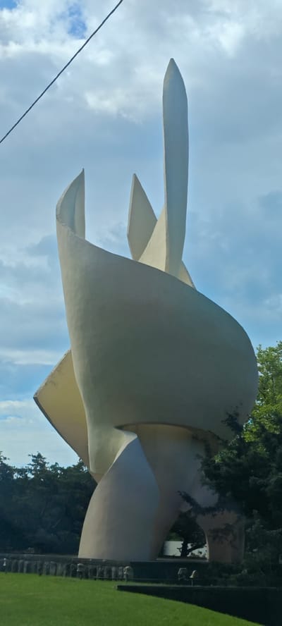 A white abstract sculpture stands tall outdoors in Novomikhaylovsky, Russia, set against a backdrop of a cloudy blue sky. The sculpture is composed of curved, pointed forms, suggesting movement and reaching upwards. Green trees and grass surround the base of the sculpture, and a low barrier or wall is partially visible. A power line crosses the upper left corner of the frame.