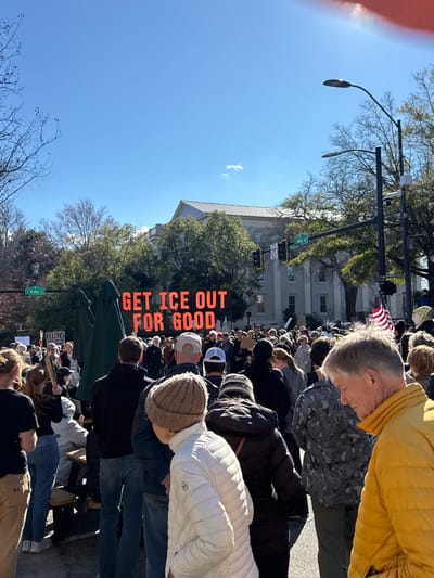 A crowded outdoor scene shows people gathered on a street. A sign reads "GET ICE OUT FOR GOOD." A building with an American flag is visible in the background, along with streetlights and trees. Many people are wearing jackets.
