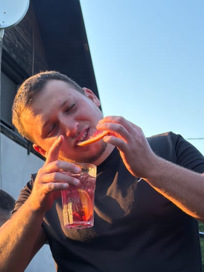In Mordarka, Poland, a young man is captured in a close-up outdoor shot. He is wearing a black t-shirt and is holding a glass of pink liquid with ice and a slice of orange. In his other hand, he is holding a slice of grapefruit to his face. The man's eyes are closed, and he appears to be enjoying the taste of the grapefruit. The lighting suggests it is late afternoon or early evening, with warm sunlight illuminating his face and arms. The background shows a portion of a building with a dark roof and a clear blue sky.