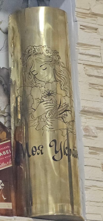 The image shows a cylindrical object, likely a piece of artillery casing, decorated with intricate line art. The artwork depicts a stylized portrait of a woman's face with long, wavy hair adorned with flowers and wheat stalks. Her hands are gently holding a bouquet of flowers and grains. The outline of Ukraine's map is subtly integrated into the design, framing the woman's face and neck. To the right of the portrait, in large, ornate Cyrillic script, is the text "Моя Україна" which translates to "My Ukraine". The object has a polished, golden sheen and rests on a surface in what appears to be an indoor setting, possibly a shelf or mantelpiece, with a textured, light-colored wall behind it. The overall impression is one of patriotic sentiment and artistic craftsmanship.