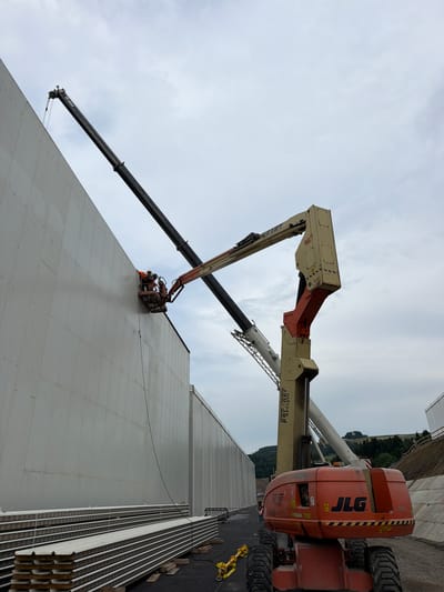 The image shows a construction scene in Skrzydlna, Poland. A large, light-colored, prefabricated building wall is under construction, with stacks of similar wall panels lying horizontally at its base. Two construction workers in orange safety vests and hard hats are visible in a cherry picker basket, working on the upper section of the wall. Above them, a dark boom crane extends over the wall. To the right of the wall, a large orange JLG aerial work platform with the number "800AJ" and "JLG" logo is positioned. In the background, a wooded hillside is visible under an overcast sky, with a sloped, engineered embankment to the right of the construction area.