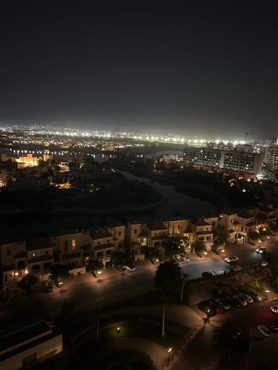 The image depicts a nighttime aerial view of a residential area with a body of water running through it. The scene is lit by artificial lights from buildings and streetlights, creating a contrast between illuminated areas and shadows.
On the right side of the image, a street is visible with several parked cars. The houses are of a uniform architectural style, with multiple stories and tiled roofs. Trees are interspersed throughout the residential area, adding to the landscape. A distinct water channel or canal runs through the middle of the scene, reflecting the lights.
On the left side of the image, a more densely built-up area is visible with taller buildings, suggesting a different part of the urban landscape or a city skyline in the distance. Lights from these buildings extend far into the background, appearing somewhat hazy, possibly due to atmospheric conditions or distance. A construction crane is faintly visible in the far distance on the left.
The time of day is clearly night, indicated by the darkness and the artificial lighting. The weather appears clear, as the lights are sharp and distinct, though some distant lights have a slight haze. The location is identified as Al Jazeera Heritage Village, United Arab Emirates, which suggests a blend of modern development with potentially older or traditional architectural elements. No people are clearly identifiable in the scene, and there is no visible text. The overall mood is one of urban nighttime ambiance, with a sense of scale provided by the aerial perspective.