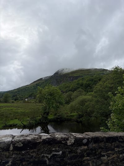 The image, when viewed in correct landscape orientation, captures a serene yet dramatic natural landscape under an overcast sky. A large, verdant mountain dominates the left side of the frame, its slopes densely covered in various shades of green trees and vegetation. The peak of the mountain is partially shrouded in low-hanging clouds or mist, creating an atmospheric and somewhat moody ambiance.

At the base of the mountain, a calm body of water, likely a river or loch, meanders through the scene, reflecting the muted light. On the distant bank, small white buildings or houses are faintly visible nestled within the green landscape. To the right, a sturdy, weathered stone wall or bridge structure occupies the foreground, its rough texture and patches of lichen evident. Beyond this wall, more of the water and distant green banks can be seen.

The sky is uniformly overcast with soft, grey clouds, indicating a cloudy daytime. No direct sunlight is visible. The overall impression is one of natural beauty and rugged tranquility, consistent with a mountainous region such as the Scottish Highlands, aligning with the provided location context of Aberfoyle, United Kingdom. No specific activity or event is taking place, and no visible text is present.