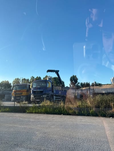 Two trucks are parked side-by-side in an outdoor lot under a clear blue sky. The truck on the left is yellow and white with "IVECO" written on the side. The truck on the right is blue and has a crane attached to its bed. Both trucks have license plates visible. A chain-link fence runs in front of the trucks, with tall dry grass and some green foliage growing behind it. In the background, there are trees and a building structure. There are reflections on the surface of what appears to be a window, showing a person's face. The scene is likely in Tsar Kaloyan, Bulgaria, given the context.
