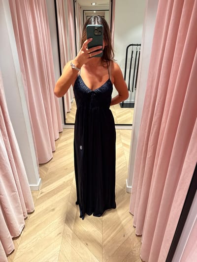 A person is depicted taking a mirrored selfie in a retail fitting room located in Las Rozas De Madrid, Spain. The individual, largely obscured by a dark green smartphone held at chest height with both hands, wears a long, dark blue sleeveless dress featuring spaghetti straps, a ruched V-neck bodice, and a small light-colored circular emblem on the left hip. A silver-toned watch or bracelet is visible on the left wrist, and a ring is on the right hand. The person's right hand rests on their hip. The fitting room features two floor-length, pleated light pink curtains framing the central mirror. The floor is light-toned wood parquet arranged in a herringbone pattern. The reflection in the mirror reveals a portion of a garment rack with dark hanging items to the right.