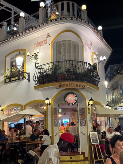 The image captures a nighttime scene on a street in Nerja, Spain, featuring the exterior of a building with multiple levels and a restaurant or bar named "Sevillano." The building's facade is white with yellow trim and arched windows. Outdoor seating areas with umbrellas and tables are set up in front of the establishment. Several people are visible, some seated at tables, others standing, and some walking by. The lighting is dim, with streetlights and the restaurant's interior illuminating the scene. The overall atmosphere appears to be lively and typical of a tourist area at night.