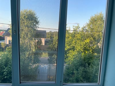 The image shows a view from a window with a screen, looking out onto a residential area. The foreground is dominated by the white window frame and the screen mesh, which slightly distorts the view of the background. The background reveals a suburban landscape with houses, fences, and abundant greenery from trees and bushes. In the distance, there are power lines and a clear blue sky with a few wispy clouds. The overall scene depicts a typical sunny day in a rural or suburban setting. The location is identified as Bilohorodka, Ukraine. There are no people or animals visible in the image. The scene appears to be a still, quiet moment captured from indoors.