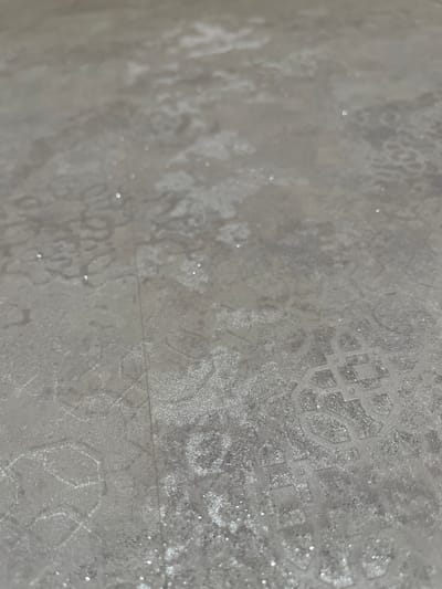 The image shows a close-up, low-angle view of a light-colored, textured surface with a repeating geometric pattern and subtle sparkle. The surface appears to be a fabric or wallpaper. There are no people or specific objects clearly identifiable in the scene. The environment is a still, indoor setting, possibly a home or commercial space. The light source is diffuse, creating soft highlights and shadows that emphasize the texture and pattern. The image is likely a detail shot of a decorative material. There is no information in the image that can be used to determine its location in the city of Keles, Uzbekistan.