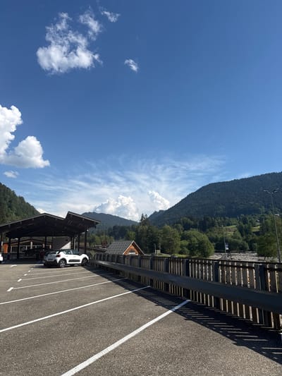 The image depicts a view from a paved parking lot in Saint-Jean-d'Aulps, France, looking towards a densely forested mountain range under a bright blue sky with scattered white clouds. In the foreground, white parking lines are clearly marked on the asphalt. A white hatchback car is parked under a wooden covered structure, which appears to be part of a building with a slanted roof. To the right of the parking lot, a wooden fence with vertical slats runs along a slight embankment. Beyond the fence, there are trees and buildings, including a structure with a sign that reads "Bienvenue." The mountain slopes are covered in evergreen trees, and a riverbed or rocky area is visible in the middle ground. Streetlights are present along the embankment. The lighting suggests it is daytime, likely late morning or early afternoon given the shadows cast by the fence.