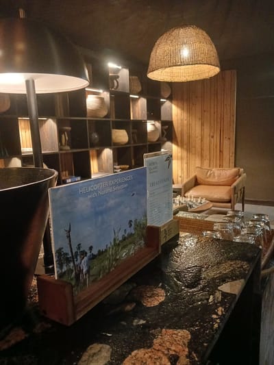 The interior shot showcases a dimly lit space, possibly in Nwanaeanoka, Botswana, featuring a counter or bar area made of a dark, textured material. Several stemmed glasses sit atop a woven tray behind a wooden display case. A brochure advertising "HELICOPTER EXPERIENCES with Natural Selection" is prominently displayed, next to another one titled "NORTH ISLAND, OKAVANGO TREATMENTS". A light fixture enclosed in a woven shade illuminates the space above shelves filled with wicker baskets and other items. A seating area with cushioned chairs is positioned near a wall adorned with wooden planks.