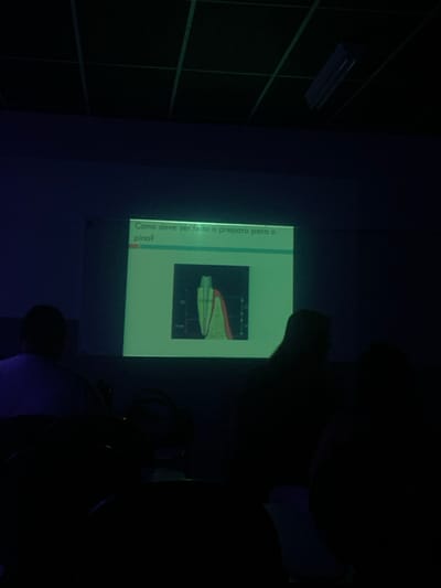 The image depicts an indoor scene in a darkened room located in Lauro de Freitas, Brazil, consistent with a lecture hall or classroom. A brightly illuminated projector screen, serving as the primary light source, is centered on a wall. The screen displays a presentation slide featuring Portuguese text: "Como deve ser feito o preparo para o pino?" (How should the preparation for the pin be done?), accompanied by a detailed anatomical diagram of a tooth's root canal system, illustrating an inserted dental post or pin with measurement indicators. In the foreground and midground, at least three human figures are visible in silhouette, seated and facing the screen, indicating their role as an audience. The ceiling above exhibits a grid pattern and includes a visible fluorescent light fixture. No active interactions among the individuals are discernible; their action is passive observation of the projected content.