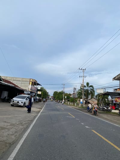 The image depicts a street scene in Cottrueng, Indonesia, under a cloudy sky. The perspective is from the side of the road, looking forward. On the left side of the road, there are buildings with corrugated metal roofs and concrete structures, one of which has a sign that reads "AKEL". A white SUV is parked on the left shoulder, and a person in dark clothing is standing near it. Further down the road, there are more buildings, palm trees, and utility poles with numerous electrical wires.
The road itself is asphalt with yellow lane markings. Several motorcycles are present. In the foreground on the right side, a person on a motorcycle is visible. Further down the road, more people on motorcycles and a yellow truck are traveling. The overall atmosphere suggests a typical day in a moderately developed area, with a mix of commercial buildings and residential structures lining the main thoroughfare.