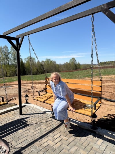 A woman sits on a swing bench in Kamennoye, Russia, on a sunny day. She wears a blue and white striped outfit and light-colored shoes. The swing bench is suspended by chains from a dark-colored wooden frame. The background includes a field and trees under a clear blue sky. A paved area with rectangular stones is visible adjacent to the swing.