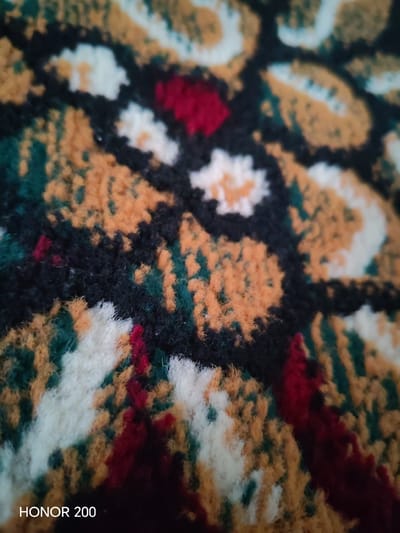 The image shows a close-up of a textured fabric, possibly a carpet or tapestry, with a repeating pattern of abstract shapes in shades of orange, yellow, white, black, and dark green. There are also small accents of red within the pattern. The lighting is somewhat dim, and the focus is soft, giving the image a slightly blurred appearance. In the lower left corner, the text "HONOR 200" is visible, indicating the device used to capture the image. There are no people, animals, or identifiable landmarks present. The image is a detailed view of a textile design and does not depict any specific scene in the city of Kakaydi, Uzbekistan, or any other urban environment.