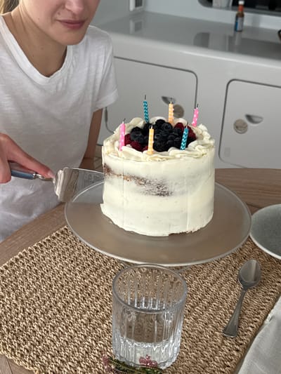 The image shows a person cutting a cake with a cake server. The cake is frosted with white icing and topped with fresh berries and birthday candles. It sits on a clear glass cake stand. A glass of water, a spoon, and a ceramic bowl are on a woven placemat next to the cake. The location is not specified.