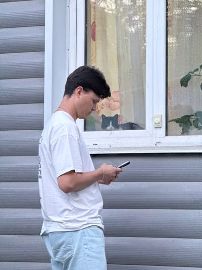 A person is standing indoors, looking at a mobile phone. The person is wearing a white t-shirt and light blue jeans. The background consists of a window and vertical blinds. The location cannot be determined as Ягул, Russia with the provided information.