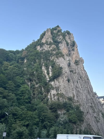 The image shows a steep, rugged mountain face covered in sparse vegetation and trees. The rock is weathered and appears to be limestone. The mountain dominates the right side of the frame, with its peak reaching towards a clear blue sky. A dense forest covers the left side of the mountain, contrasting with the barren rock face. In the foreground, a portion of a white van is visible, along with a black lamppost. The overall scene suggests a natural landscape, possibly a natural park or a mountainous region near a settlement. The presence of the van and lamppost indicates that this is a populated area, likely on the outskirts of Tengealti, Azerbaijan, known for its mountainous terrain.