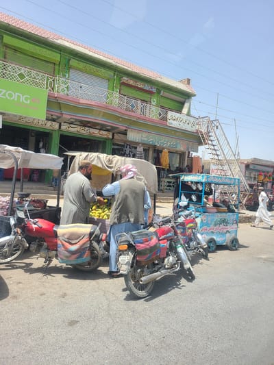 The image depicts a street scene in Saranan, Pakistan, likely during the daytime under clear skies. The primary subjects are two men and several motorcycles parked on a paved road. One man, wearing a light-colored shalwar kameez and a cap, is standing and appears to be interacting with a stall. The other man, wearing a similar outfit and a checkered headscarf, is partially visible, also engaged with the stall.

The stall itself is a colorful, cart-like structure, painted blue and adorned with text, possibly a vendor's advertisement. It is laden with various items, including what appear to be mugs or glasses, and a selection of fruits, possibly bananas, are displayed prominently. Several motorcycles, some red and others silver, are parked adjacent to the stall. These vehicles are decorated with colorful saddlebags or cushions.

Behind the stall and motorcycles, a building with green and pink trim is visible. This building features windows and decorative elements, with some signage also present. A prominent green sign with "ZONG 4G LET'S GET DIGITAL" is attached to the building, indicating a mobile network provider. The overall setting appears to be a bustling street or marketplace, with evidence of commercial activity. The image is shot from a slightly angled perspective, with the scene oriented vertically. The presence of overhead power lines and the bright sunlight suggest an outdoor, everyday scenario. The text within the image includes Arabic script, phone numbers, and brand names.