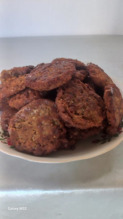A close-up shot shows a bowl filled with golden-brown, irregularly shaped patties. The patties appear to be fried and have a textured surface, with some appearing slightly crispy around the edges. The bowl itself is off-white and has a decorative rim with a pattern of red and green floral motifs. The patties are piled high in the bowl, filling most of the frame. The background is a light-colored surface with a subtle embossed pattern, possibly a tablecloth. To the right of the bowl, the text "Galaxy M32" is visible vertically, indicating the device used to capture the image. The lighting suggests an indoor setting, and the overall impression is one of a home-cooked meal. Given the location context of Ынтымак, Kyrgyzstan, the patties could be a local dish, such as kutub or meat patties.