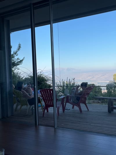 The scene depicts an interior view through large sliding glass doors onto an outdoor wooden deck in the Merom HaGalil Regional Council, Israel, with a panoramic landscape visible beyond.
In the immediate foreground, a dark wooden floor is partially visible, indicating an indoor living space. The large glass panels reflect ambient light.
On the outdoor deck, constructed of horizontal wooden slats, three to four individuals are present. On the left side, a woman in a dark top is seated in a yellow plastic chair, her profile visible as she appears to be conversing with another person largely obscured behind her. Near her on the deck is a silver insulated bottle. Toward the center, a red plastic Adirondack-style chair is positioned, next to which a small, multi-colored table is partially visible. Further to the right, an individual, possibly female, wearing a light-colored top and shorts, is seated in another red plastic Adirondack-style chair, looking down at an object held in their hands. A pair of black flip-flops are on the deck in front of this chair. On the far right, a rustic wooden bench is situated.
Beyond the deck, a dense layer of green vegetation, including trees and bushes, descends the hillside. In the middle distance, a vast valley or plain stretches out, displaying a hazy, muted landscape. In the far background, under a clear, bright blue sky, a prominent mountain range, consistent with Mount Hermon, is visible. The scene is illuminated by natural daylight, suggesting late afternoon or early evening.