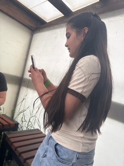 A young girl with long, dark hair pulled into a ponytail is holding a smartphone in her hands, looking down at it. She is wearing a light-colored t-shirt with dark trim on the collar and sleeves, and blue jeans. A green wristband is visible on her left wrist. She is standing or sitting near wooden benches, with tall grass in the background. The environment appears to be an outdoor structure with a translucent roof and walls. The image was taken in Zaliznychne, Ukraine.