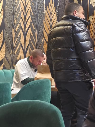 Two men are captured in the image. One, wearing a black puffer jacket and dark pants, stands near a wall adorned with a leaf pattern. The other man is seated on a green chair, sporting a white shirt. The green chair occupies a substantial portion of the foreground. The image is shot indoors with neutral lighting.