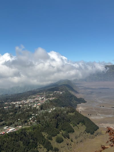 The image shows a panoramic view from a high vantage point overlooking the village of Cemorolawang, Indonesia, nestled on a hillside. The foreground is dominated by a steep, tree-covered slope, with a cluster of buildings appearing on the left side of the slope, some with red roofs. A town or village spreads across the middle ground, characterized by numerous houses with red, orange, and gray roofs, interspersed with trees. A red and white communication tower is visible within the village. The terrain slopes downwards from the village into a vast, arid plain in the background, marked by sandy soil and meandering tracks. The plain is bordered by distant, hazy mountains and hills. The sky above is a clear, bright blue, with scattered white clouds congregating along the horizon, partially obscuring the distant mountain ranges. The overall scene depicts a rural settlement situated between lush, green hillsides and a stark, open plain under a bright sky.