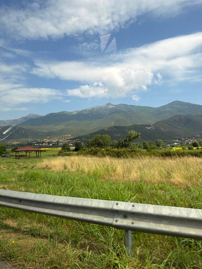 The image captures a rural landscape on a bright, partly cloudy day, with a highway guardrail prominently in the foreground. Beyond the guardrail, a field of tall green and yellow grass extends towards a cluster of trees and a red-roofed gazebo. In the middle ground, a small village is visible nestled against the foothills of a large, verdant mountain range. The mountains dominate the background, their slopes covered in dense vegetation, disappearing into a hazy sky. The scene appears to be set in or near Μέλισσα, Greece, given the distinctive mountain topography and rural setting often associated with the region. There are no people or animals visible in the frame, and the overall impression is one of natural beauty and peacefulness.