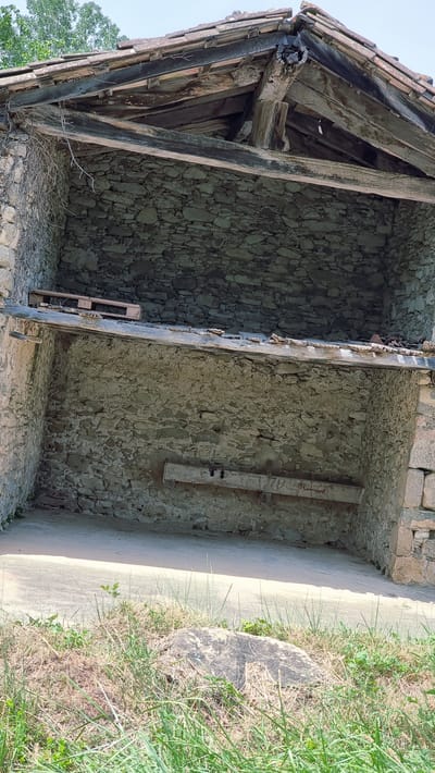 The image shows the interior of a partially collapsed stone structure, likely an old barn or outbuilding, located in the municipality of La Vall d'en Bas, Spain. The structure's roof is missing in places, revealing exposed wooden beams and roof tiles. The stone walls are rough and uneven, characteristic of traditional rural construction. Inside, a wooden pallet rests on a horizontal wooden beam in the upper section. Another wooden beam is affixed horizontally to the lower wall. The ground inside the structure is a concrete or packed earth surface, and the exterior shows overgrown grass and a large rock in the foreground. The scene suggests abandonment and decay, typical of historical rural architecture undergoing natural deterioration. There are no people or animals visible in the image.