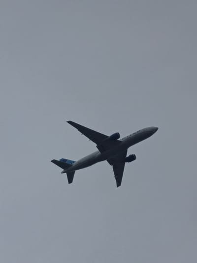 An airplane is captured from below against a hazy, overcast sky. The aircraft, identified as a United Airlines plane by the visible "UNITED" text on its fuselage and blue accents on its tail, is in mid-flight, its wings and engines clearly defined against the pale backdrop. The plane is angled upwards and to the right, suggesting it is climbing or cruising. The image appears to be taken looking directly up into the sky, with no other elements of the surrounding environment, such as buildings or landscape, visible. The overall impression is one of a solitary aircraft navigating a muted, featureless sky.