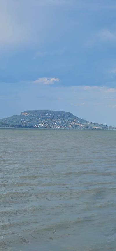 A wide shot captures a large, flat-topped mountain with a village spread across its slopes, rising from the calm, gray waters of Lake Balaton. The mountain is covered in dense green foliage, with the village appearing as a collection of white and light-colored buildings nestled amongst the trees. The lake extends across the foreground, its surface rippled by a gentle breeze. Above the mountain, a partly cloudy blue sky dominates the upper portion of the frame, with a few wispy clouds present. The scene suggests a serene, natural landscape with human settlement integrated into the environment. This is likely the Badacsony mountain, a prominent geological feature and wine-producing region on the northern shore of Lake Balaton, near the town of Balatongyörök, Hungary.