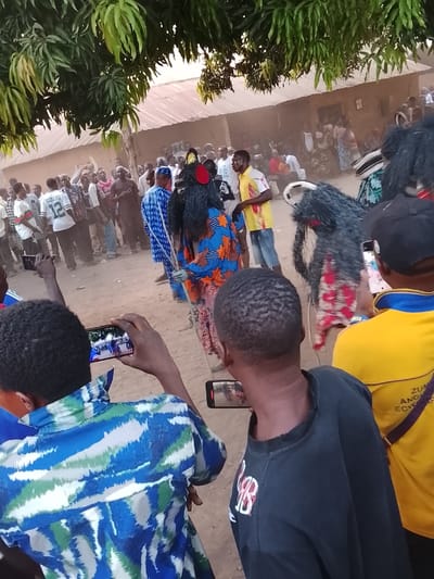 This image depicts an outdoor gathering in Tabu, Nigeria. In the foreground, two individuals are visible from behind, each holding a black smartphone horizontally to record the scene. The person on the left wears a blue, green, and white patterned shirt. The person on the right, partially visible, wears a black shirt; another individual in a yellow and blue shirt is beside them.
The middle ground features two costumed figures, identified as masquerades, surrounded by a dense crowd. The masquerade on the left has a dark, shaggy headpiece with red and yellow elements, wearing a blue, orange, and white patterned outfit, and holds a thin, light-colored staff. The masquerade on the right has a similar dark, shaggy headpiece with white accents and red fabric patches on its body. Numerous spectators, primarily men, dressed in a variety of casual clothing, including t-shirts and traditional attire, observe the central figures. One man in the crowd wears a blue patterned outfit, and another wears a white t-shirt with "RODGERS 12" printed on it.
The background shows single-story buildings with light-colored walls and corrugated metal roofs under a bright sky, partially obscured by green tree foliage overhead and what appears to be dust or haze in the air. The ground is unpaved and dusty.